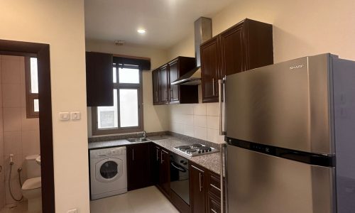 Modern kitchen with dark wood cabinets, stainless steel appliances including a refrigerator and oven, and a washing machine. A window provides natural light. A door to the left leads to a bathroom.