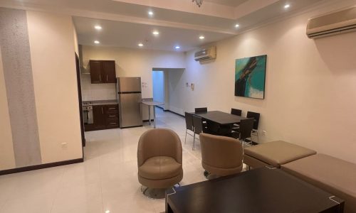 A modern, well-lit living room with a dining area and a kitchen in the background. The room features a chandelier, brown furniture, and wall art. The kitchen includes stainless steel appliances.