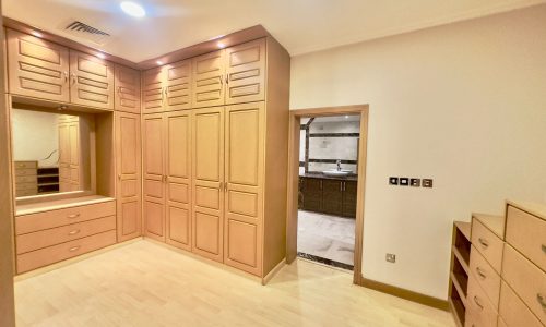 A 2 bedrooms villa in Al Janabiyah features a room with wooden built-in wardrobes and drawers. There is a large mirror with a vanity table below. An open door leads to a marble-tiled bathroom in the background. This charming residence is situated within an exclusive compound for added privacy.