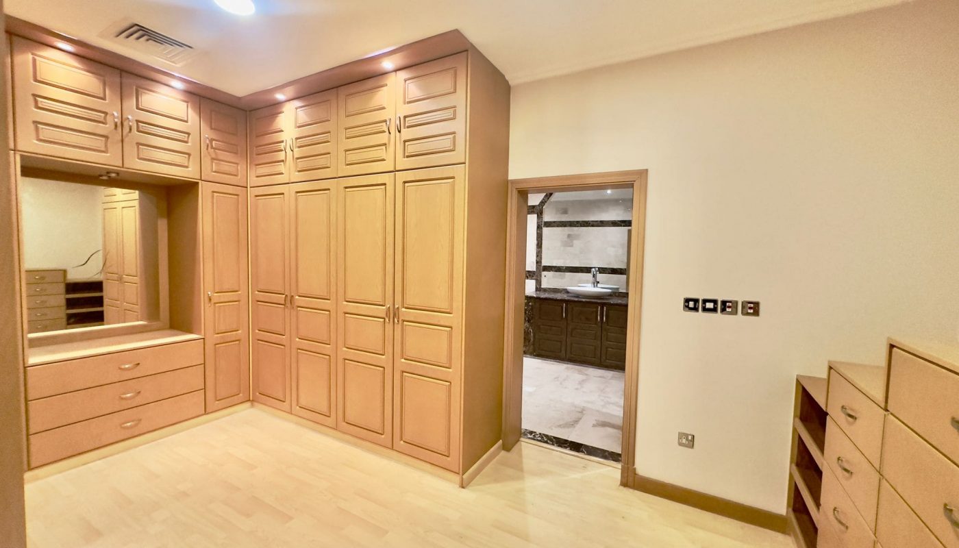 A 2 bedrooms villa in Al Janabiyah features a room with wooden built-in wardrobes and drawers. There is a large mirror with a vanity table below. An open door leads to a marble-tiled bathroom in the background. This charming residence is situated within an exclusive compound for added privacy.