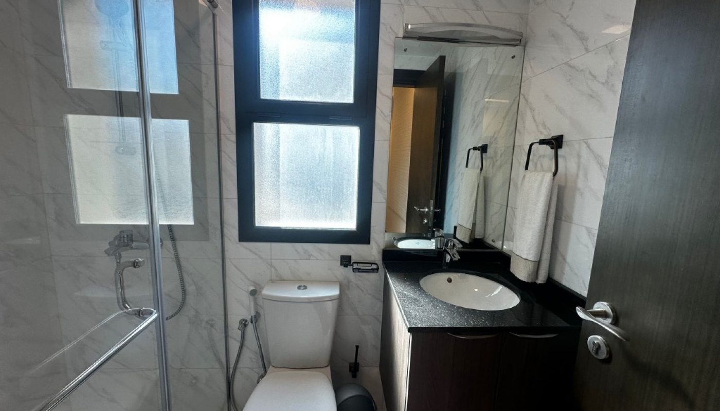 Small bathroom with a toilet, shower area with glass door, black countertop sink, mirror, and frosted window. Walls have white marble pattern tiles.