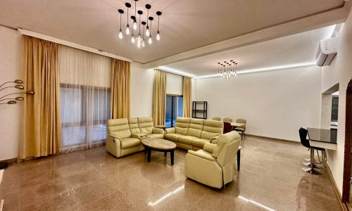 A fully-furnished living room with beige furniture and a chandelier.