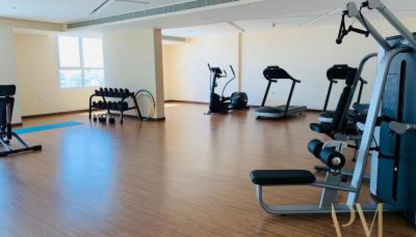 A spacious gym room with wood flooring, featuring treadmills, an elliptical machine, a stationary bike, free weights, and a multi-station weight machine.