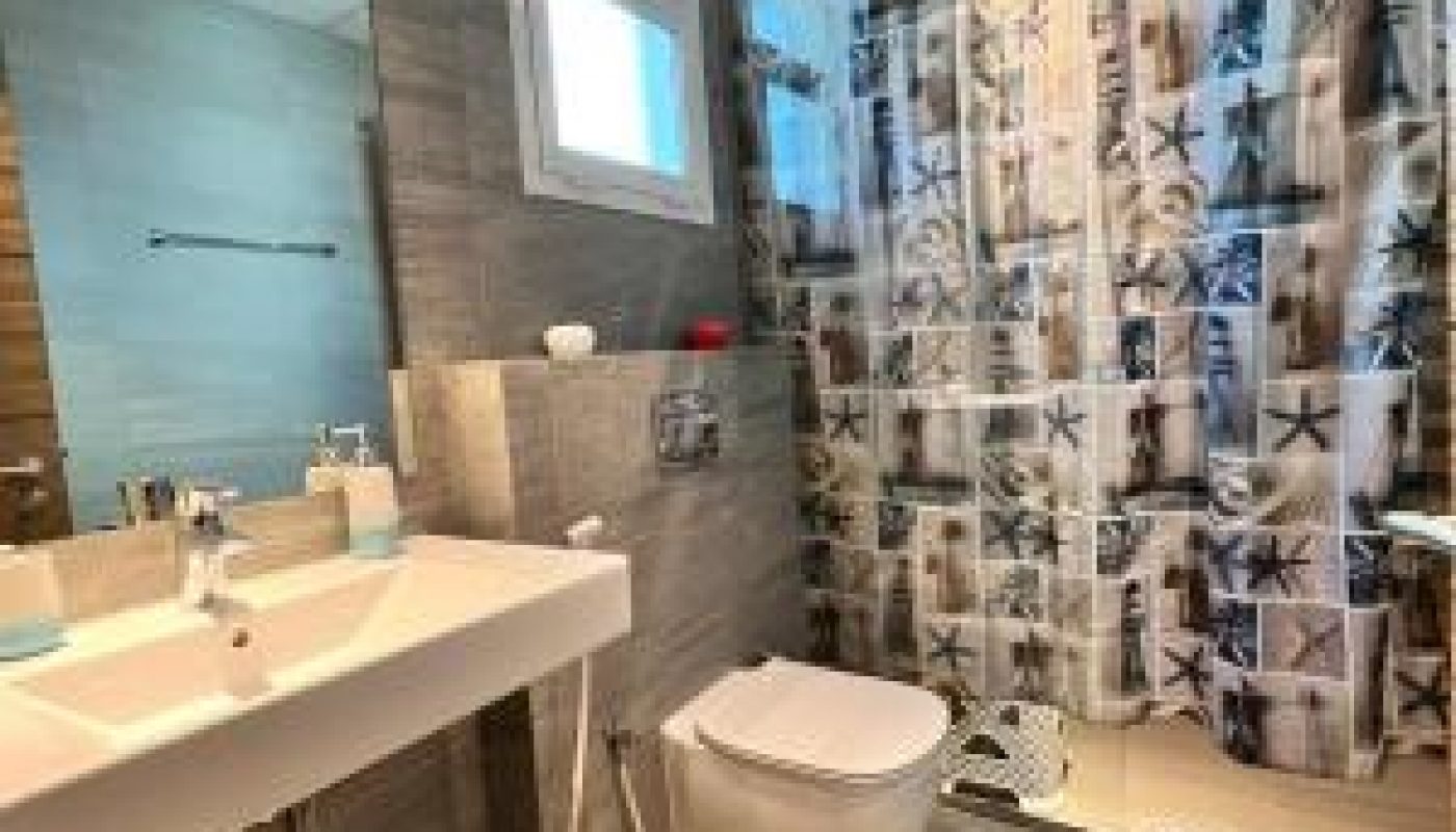 Modern bathroom with a wall-mounted sink and toilet, patterned shower curtain with sea motifs, window, and gray tile walls and floor.