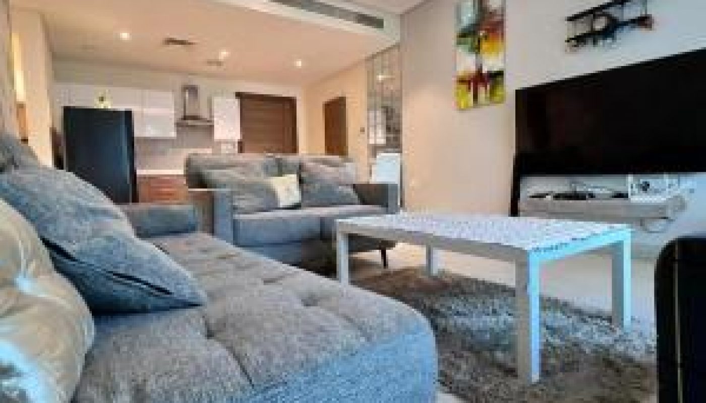 Modern living room with grey sofas, a white coffee table, a flat-screen TV, abstract wall art, and an open view into the kitchen and dining area.