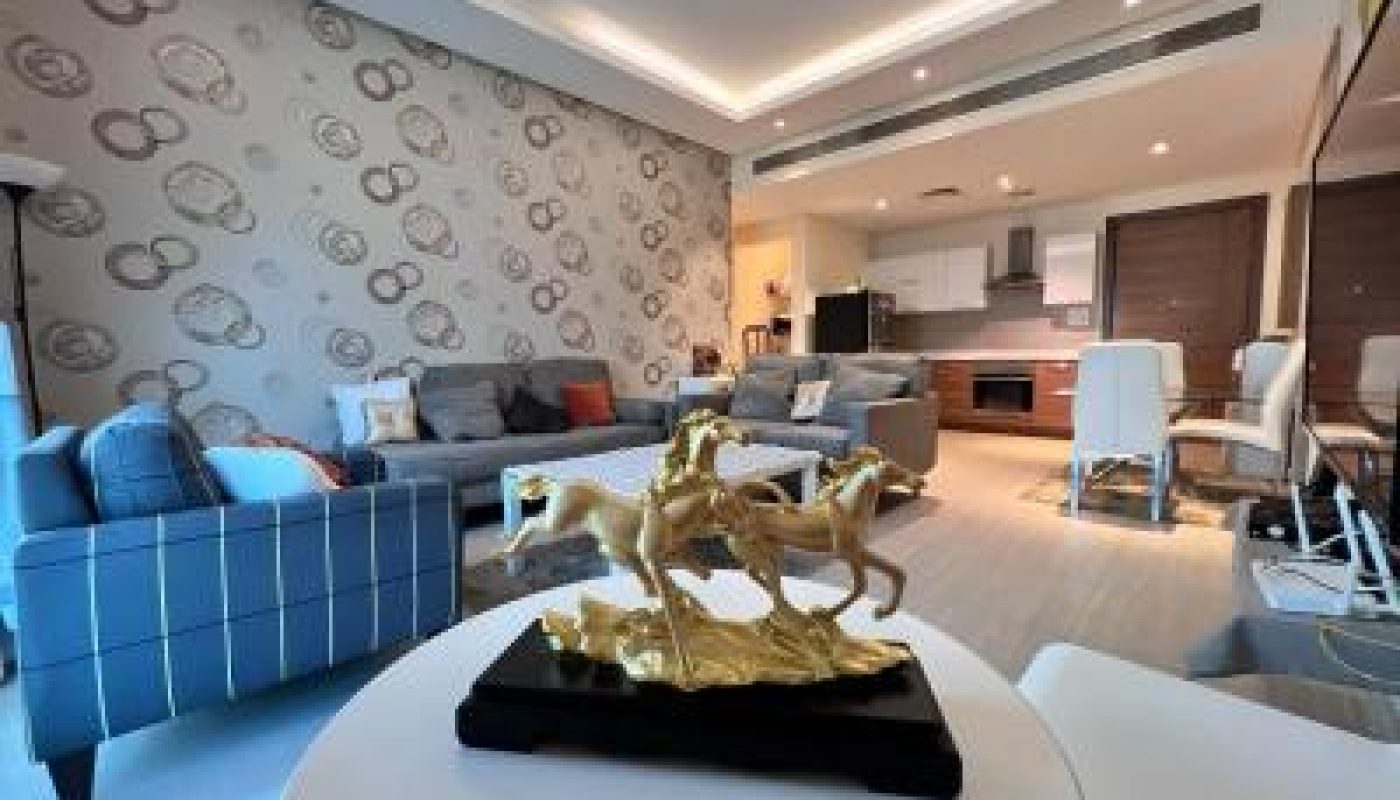 Modern living room with patterned accent wall, gray sofas, and a gold horse sculpture on a table in the foreground; open kitchen and dining area in the background.