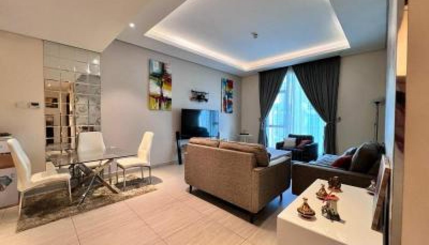 Modern living room with neutral-toned furniture, a glass dining table, wall art, large windows with dark curtains, and a TV mounted on the wall.
