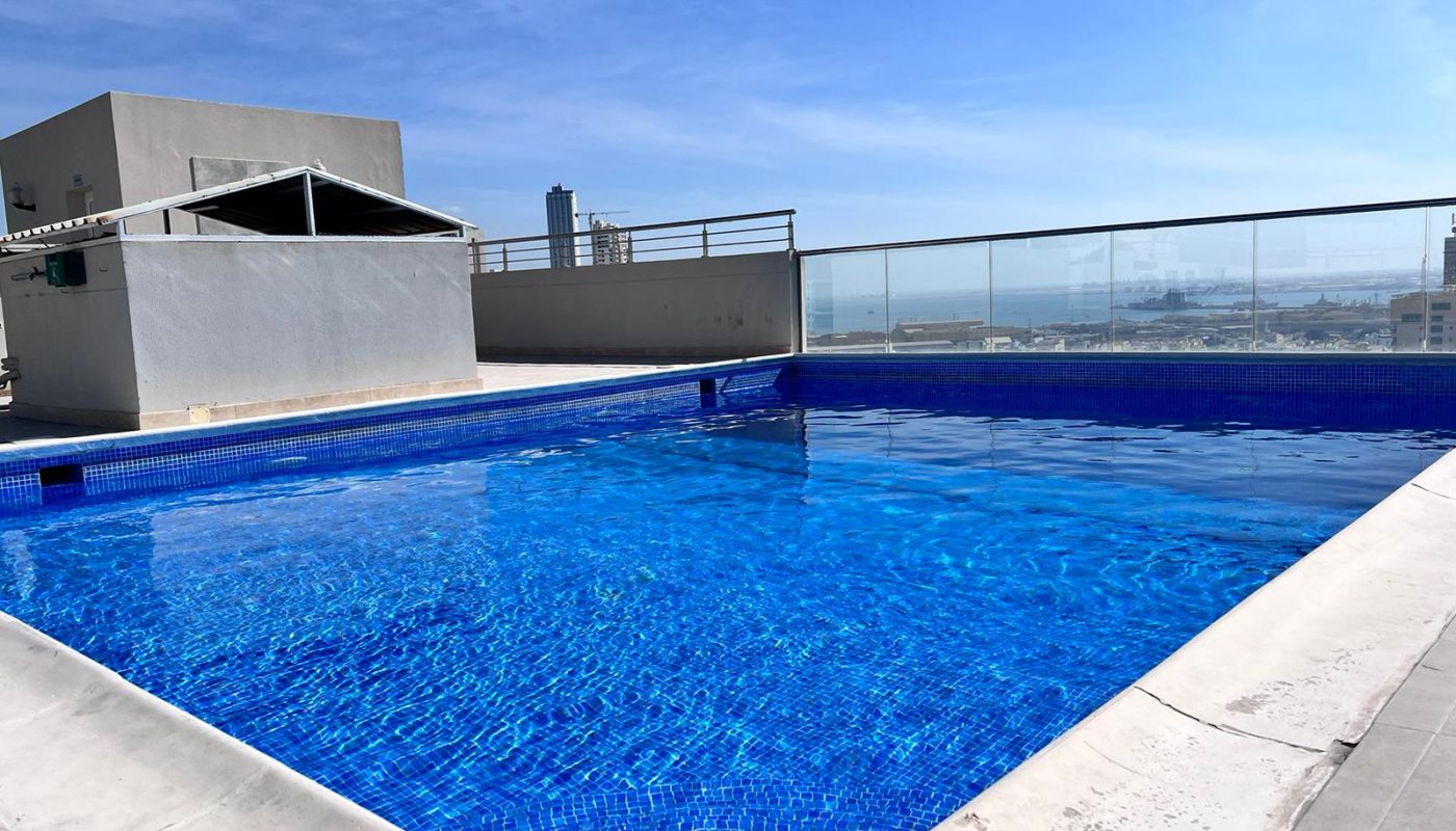 Rooftop swimming pool with clear blue water, surrounded by a white deck and glass railing, overlooking a cityscape under a clear sky.