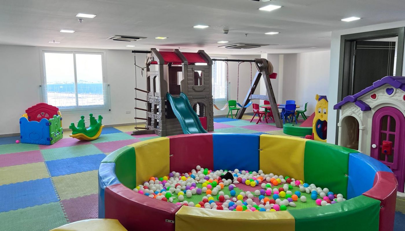 Indoor children’s playroom with colorful foam mats, a ball pit, playhouses, slides, toys, and ample natural light from large windows.