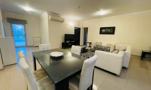 A spacious living room with a dining table and chairs in a 2BR apartment with a balcony, located in Juffair.