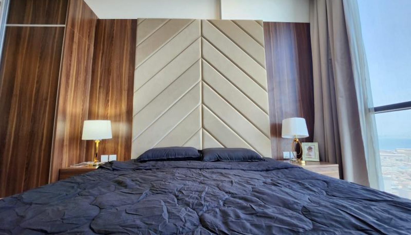 A modern bedroom with a large bed covered in a black comforter, two bedside tables with lamps, and a tall padded headboard with geometric patterns.