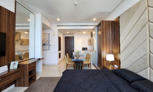 Modern studio apartment interior with a neatly made bed in the foreground, a dining area, and a kitchen visible in the background, featuring warm lighting and contemporary decor. This rental property is designed for comfort and