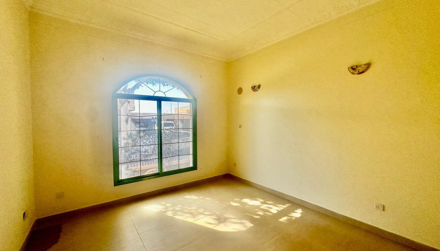 An empty room with beige walls, a large arched window, tile flooring, and two wall-mounted light fixtures sits in the semi-furnished 3 bedrooms villa. Sunlight streaming through the window casts shadows on the floor in this serene Janbyah area home.