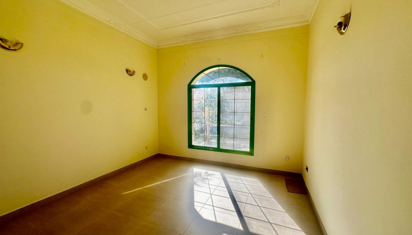 An empty room with cream-colored walls, a tiled floor, and an arched window allowing sunlight to enter. Wall-mounted lights are on both sides of the window in this semi-furnished villa located in the Janbyah area.