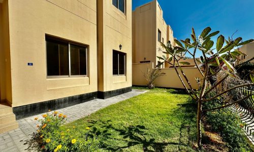 A beige villa with large windows overlooks a small, neatly maintained garden with green grass, flowering plants, and a bare tree. Located in Janbiyah, this beautiful property offers 4 bedrooms. Above, the sky is clear and blue.