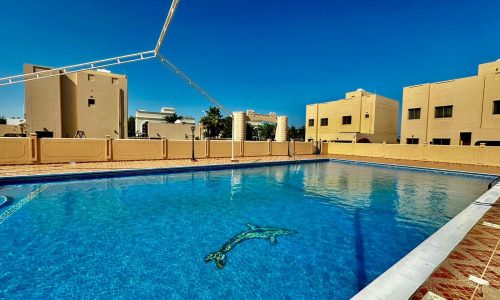 Outdoor swimming pool surrounded by beige buildings under a clear blue sky, with a dolphin design at the bottom of the pool in this stunning 4-bedroom villa in Janbiyah.