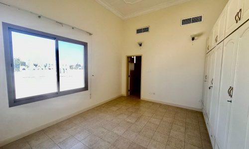 A vacant room with a tiled floor, a large window on the left wall, white built-in wardrobes on the right, and a door in the middle of the back wall sits within this 4 bedrooms villa compound in Janbiyah area. Ceiling lights and vents are also visible.