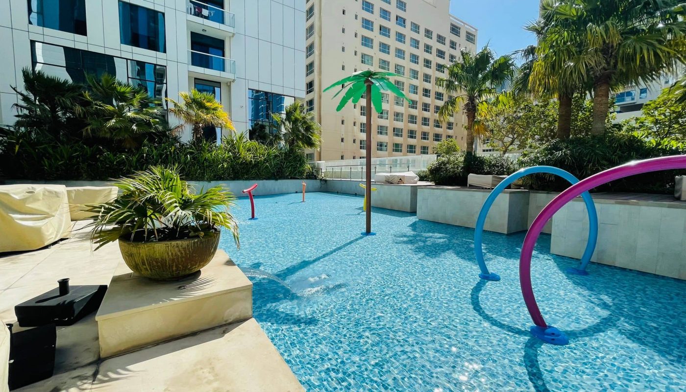 A sunlit, outdoor children's pool area with colorful, playful water features surrounded by modern buildings and tropical vegetation is part of the luxury 2BR apartment for rent in Juffair.