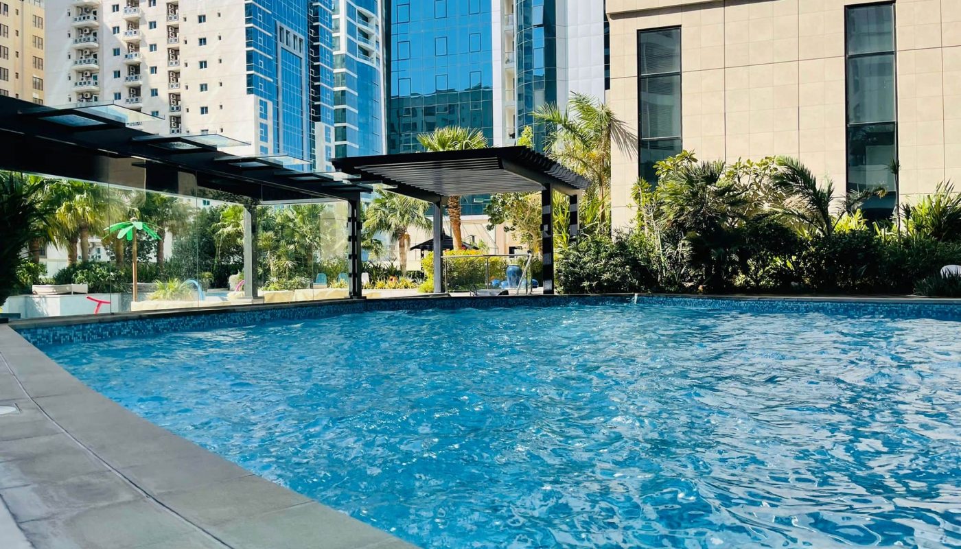 Swimming pool surrounded by modern buildings and lush greenery on a sunny day, near a luxury 2BR apartment available for rent in Juffair.