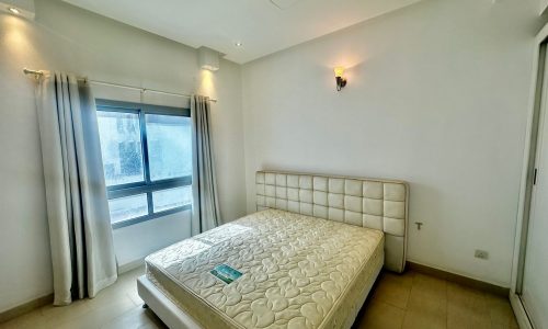 A minimally furnished bedroom with a large window, white walls, a double bed with a patterned mattress, and white curtains.