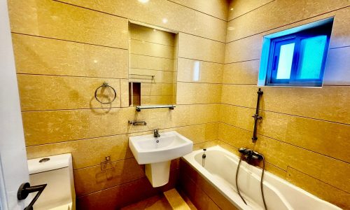 Modern bathroom with light beige tiles, a wall-mounted sink, toilet, bathtub with a shower attachment, rectangular mirror, and a small window.