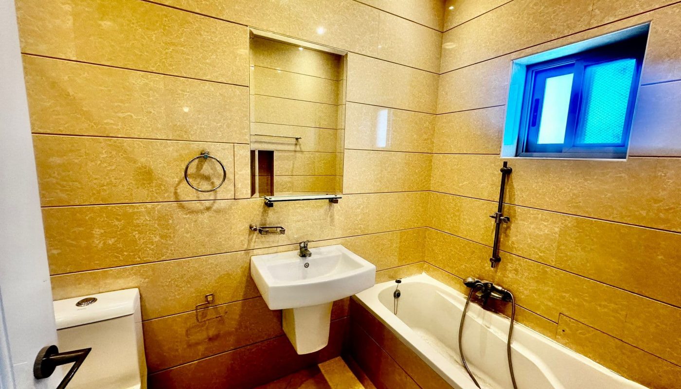 Modern bathroom with light beige tiles, a wall-mounted sink, toilet, bathtub with a shower attachment, rectangular mirror, and a small window.