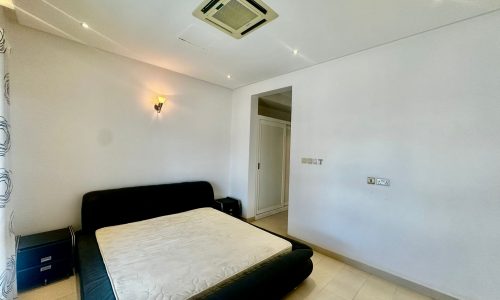 Minimalist bedroom with a black bed frame and mattress, bedside table with drawers, wall-mounted lamp, and an air conditioning unit on the ceiling. White walls and tiled floor. Closet visible in corner.