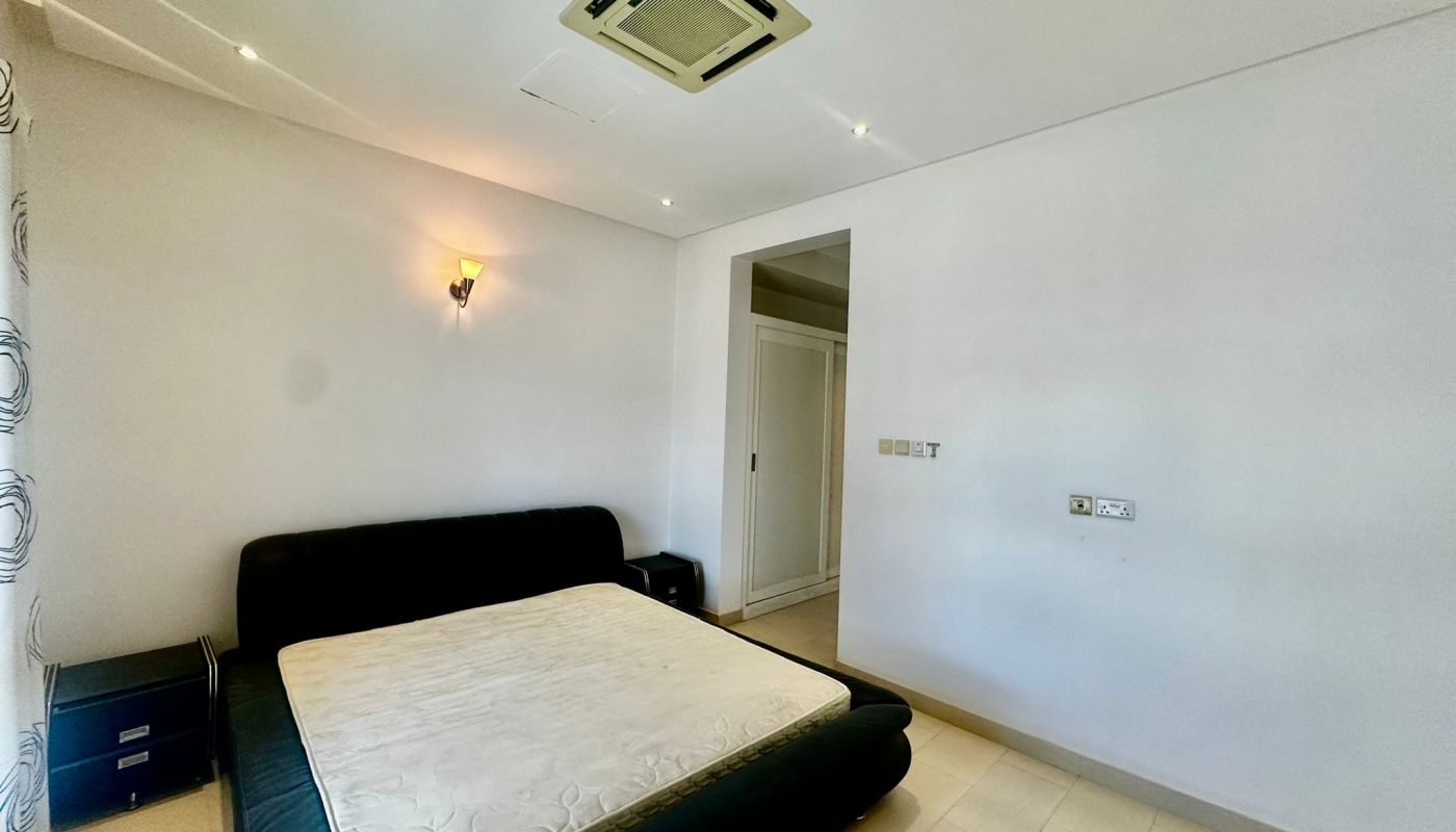 Minimalist bedroom with a black bed frame and mattress, bedside table with drawers, wall-mounted lamp, and an air conditioning unit on the ceiling. White walls and tiled floor. Closet visible in corner.