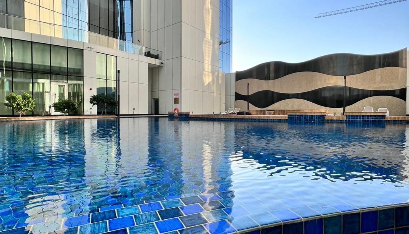 Outdoor swimming pool with blue tiles and white stones, surrounded by modern glass buildings under a clear sky.