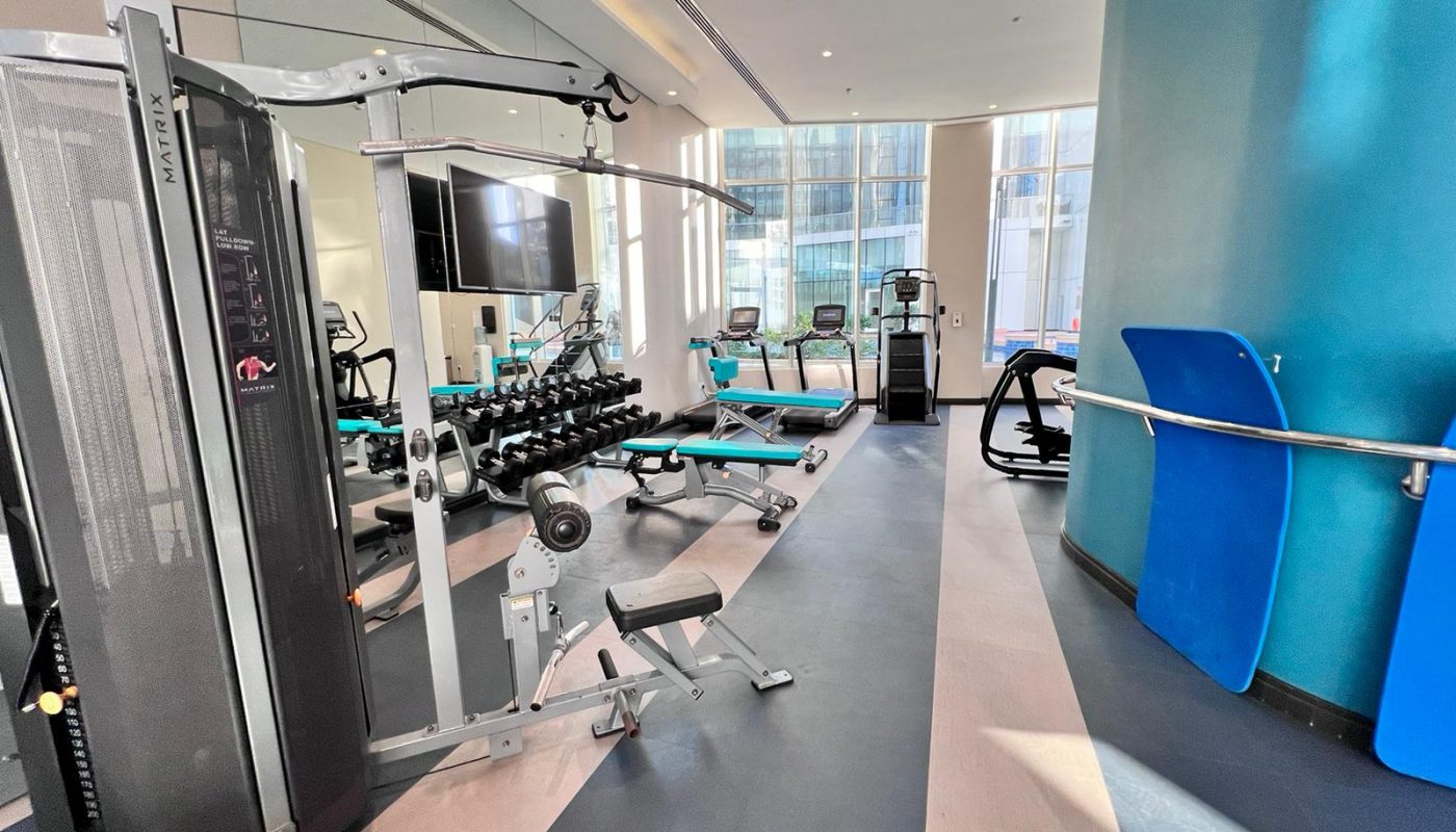 Modern gym with weight machines, free weights, benches, treadmills, and large windows letting in natural light. Blue and grey decor, mirrors line one wall.
