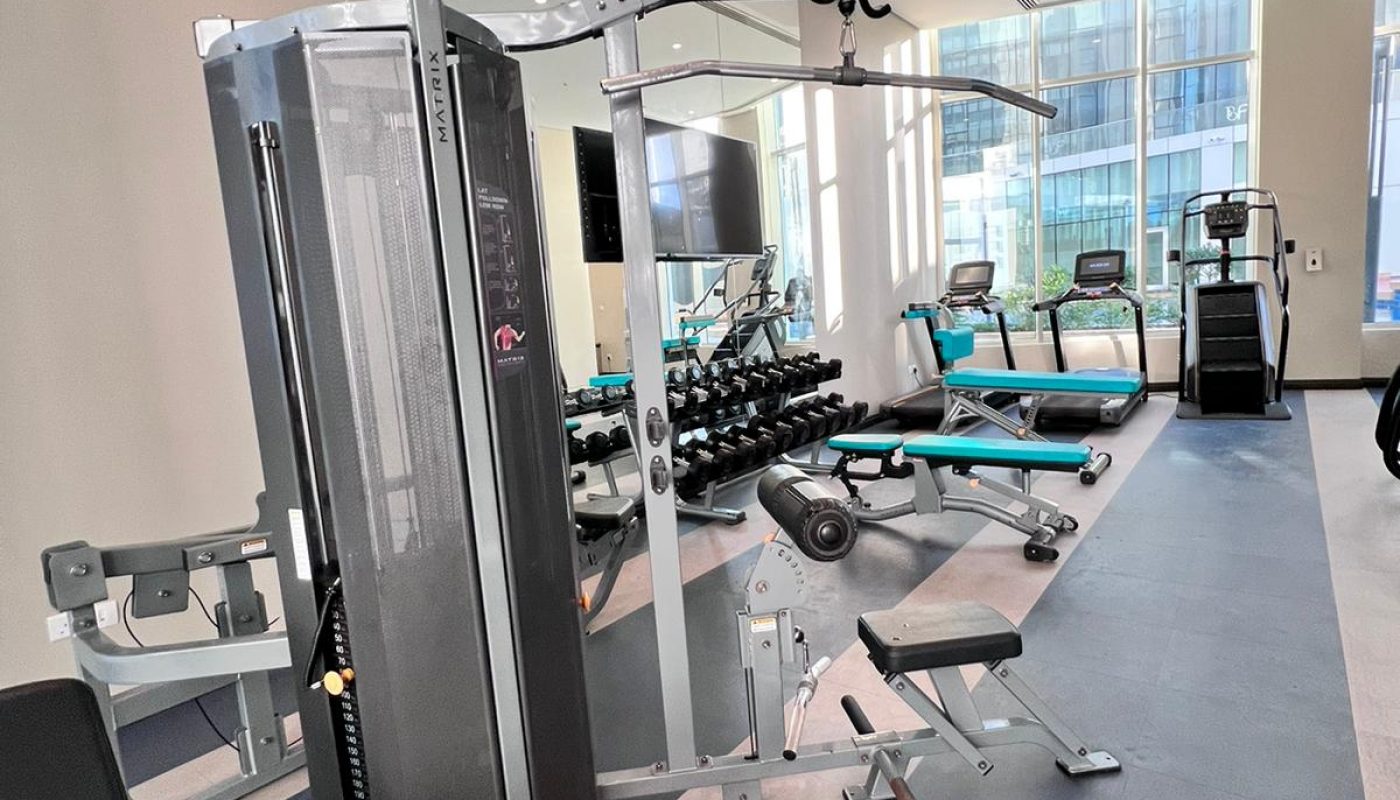 Modern gym room with weight machines, free weights, treadmills, stationary bikes, and large windows offering city views.