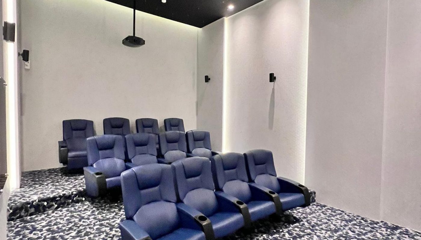 Small private theater room with two rows of blue recliner seats, patterned carpet, white walls, and a ceiling-mounted projector.