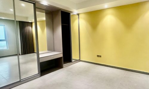 A vacant room with light yellow walls, a built-in wardrobe with mirrored sliding doors, and overhead lighting. There is a large window with dark curtains and a small built-in desk near the wardrobe.