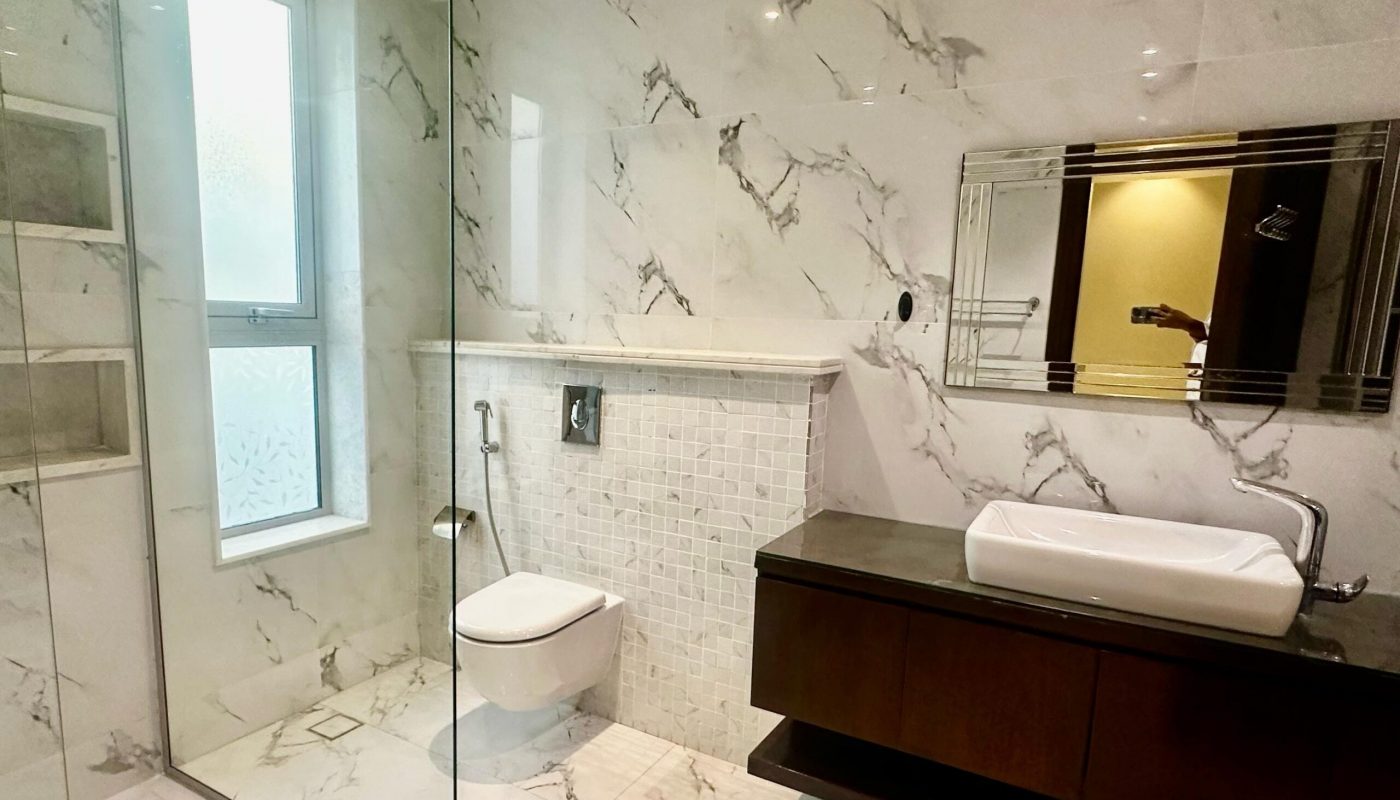 A modern bathroom with a glass-enclosed shower, wall-mounted toilet, large mirror, vessel sink, and marble tile walls and floors.