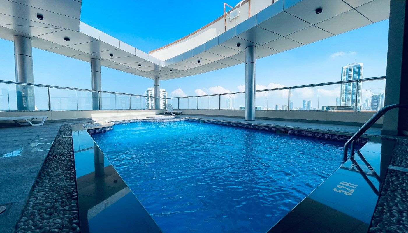 Rooftop swimming pool with clear blue water, surrounded by modern glass railings and buildings visible in the background under a bright sky.