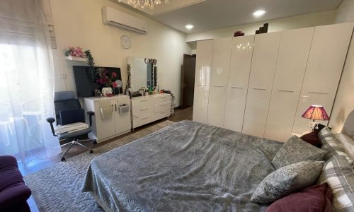 A furnished bedroom in Danat Isa Town apartment, equipped with a bed, dresser, and TV.