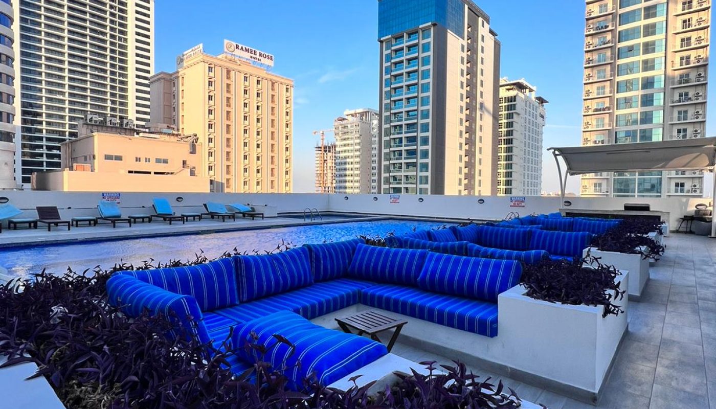 Rooftop seating area with blue-striped couches and planters, surrounded by tall city buildings under a clear blue sky.