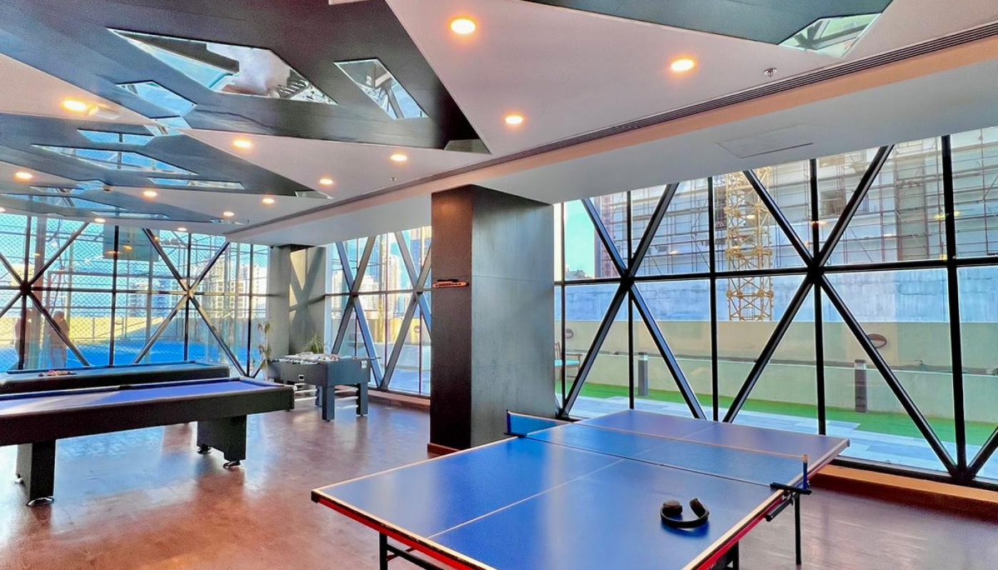 A modern game room with a ping pong table, a pool table, and large windows allowing natural light, featuring a geometric ceiling design.