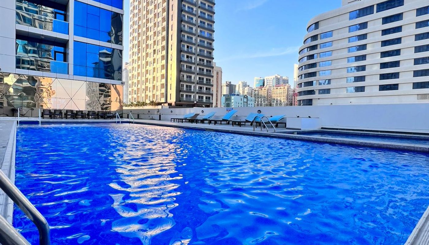 Outdoor swimming pool surrounded by modern high-rise buildings under a clear blue sky, with pool ladders visible in the foreground.