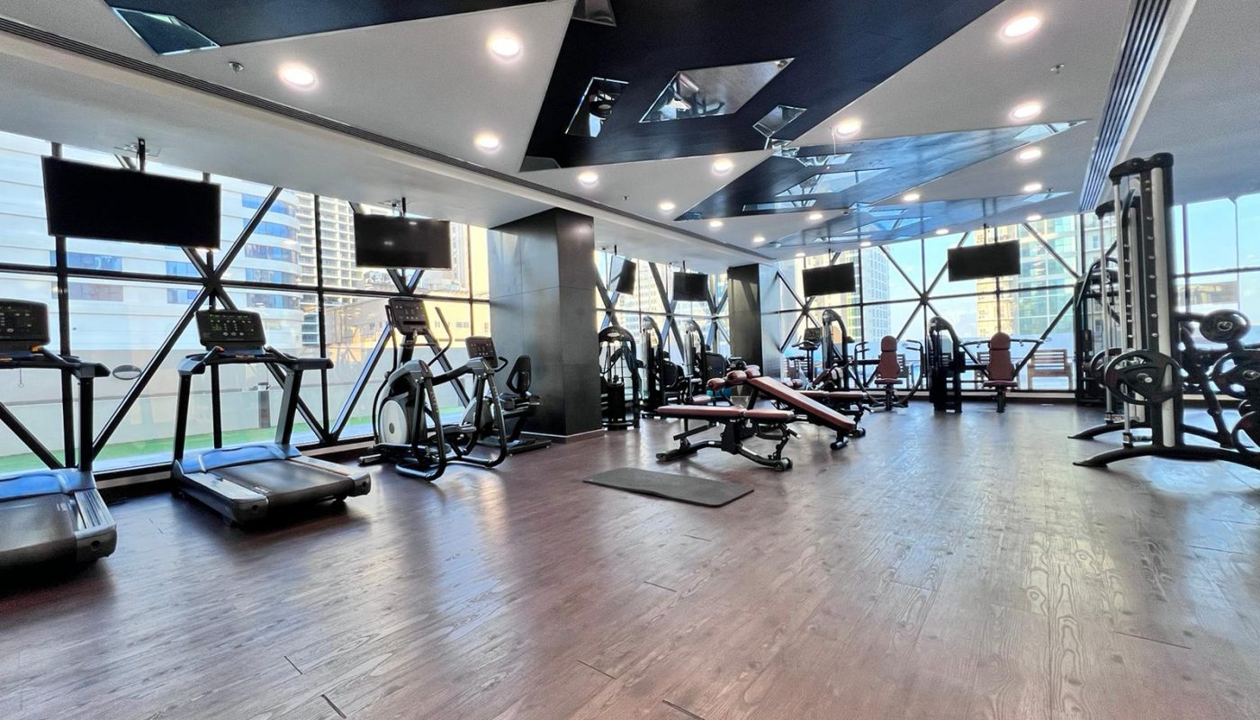 Spacious gym with treadmills, elliptical machines, weight benches, free weights, and large windows letting in natural light.