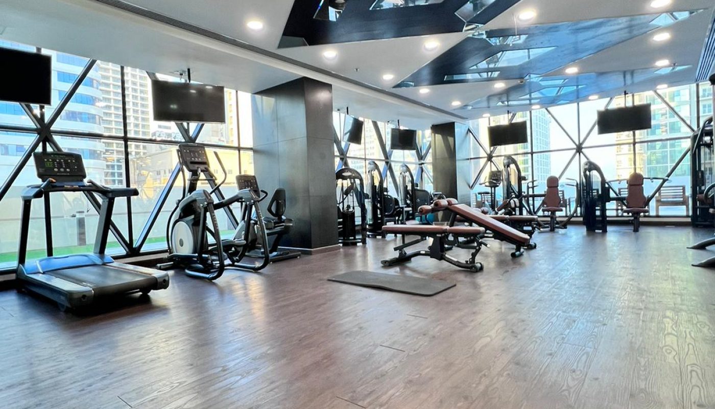 Modern gym with treadmills, stationary bikes, weight benches, and large windows overlooking city buildings. Wood flooring and ceiling lights are visible.