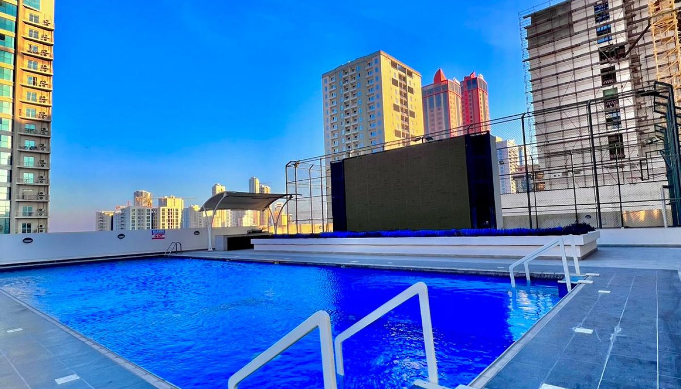 A rooftop swimming pool with blue water, surrounded by a tiled deck; high-rise buildings and construction scaffolding are visible in the background under a clear blue sky.