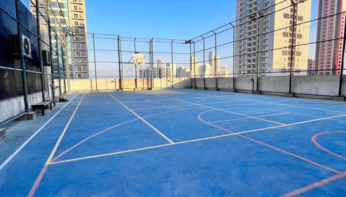 A rooftop basketball court with blue flooring, surrounded by a wire fence, with tall buildings and a clear blue sky in the background.