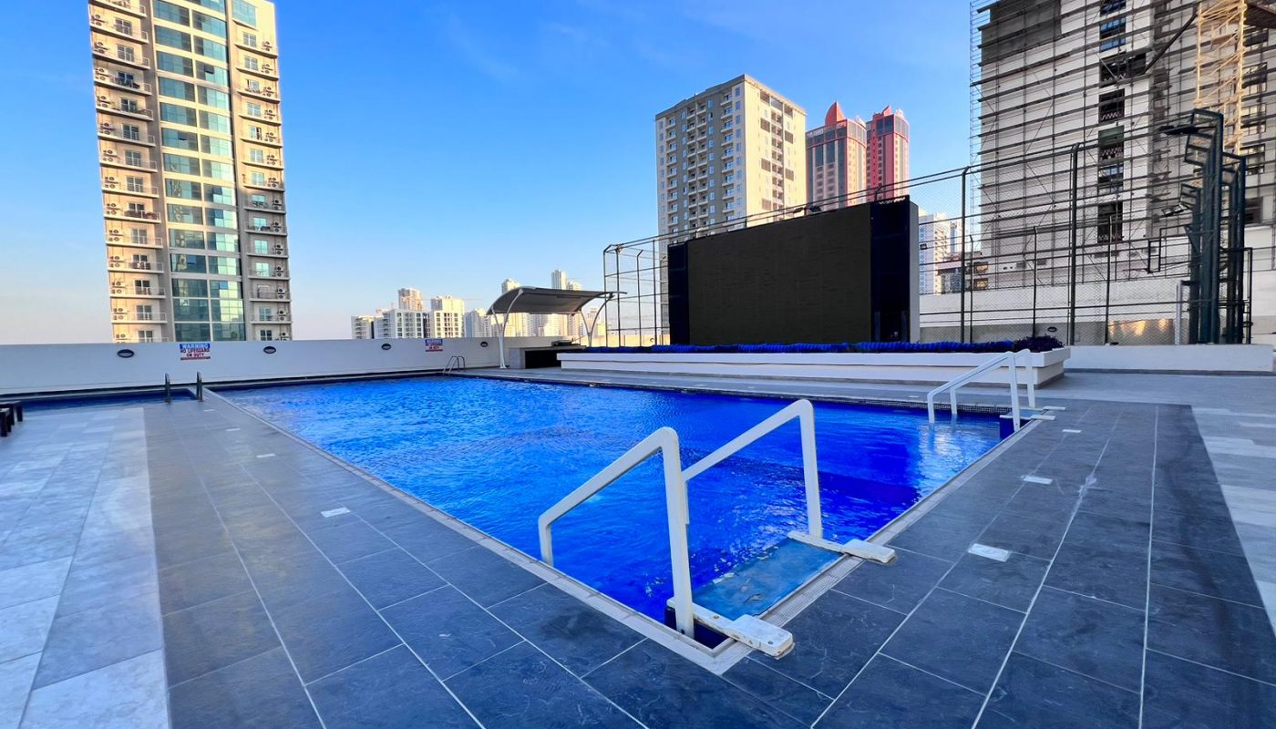 Rooftop swimming pool with blue water, surrounded by tall buildings under a clear sky, featuring a tiled deck and metal handrails.