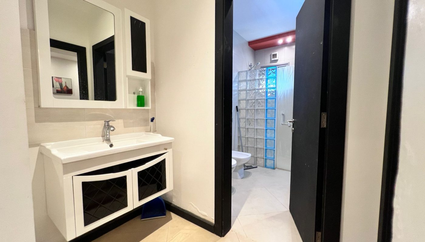 Modern bathroom with white sink and cabinet, wall mirror, soap dispenser, and a door leading to a toilet and shower area with glass block wall and tile flooring.