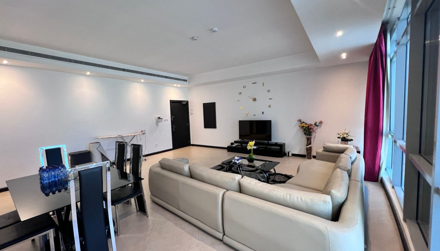 Spacious modern living room with a large beige sectional sofa, glass dining table with black chairs, TV, wall clock, and floor-to-ceiling windows with pink curtains.