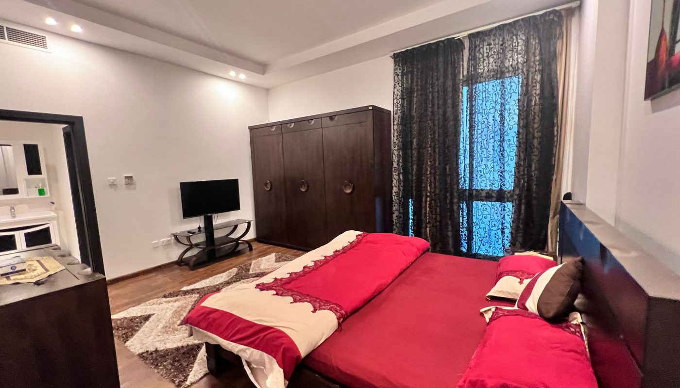 A bedroom with a double bed covered in red linens, a dark wooden wardrobe, a TV on a stand, a patterned rug, and a window with dark curtains.