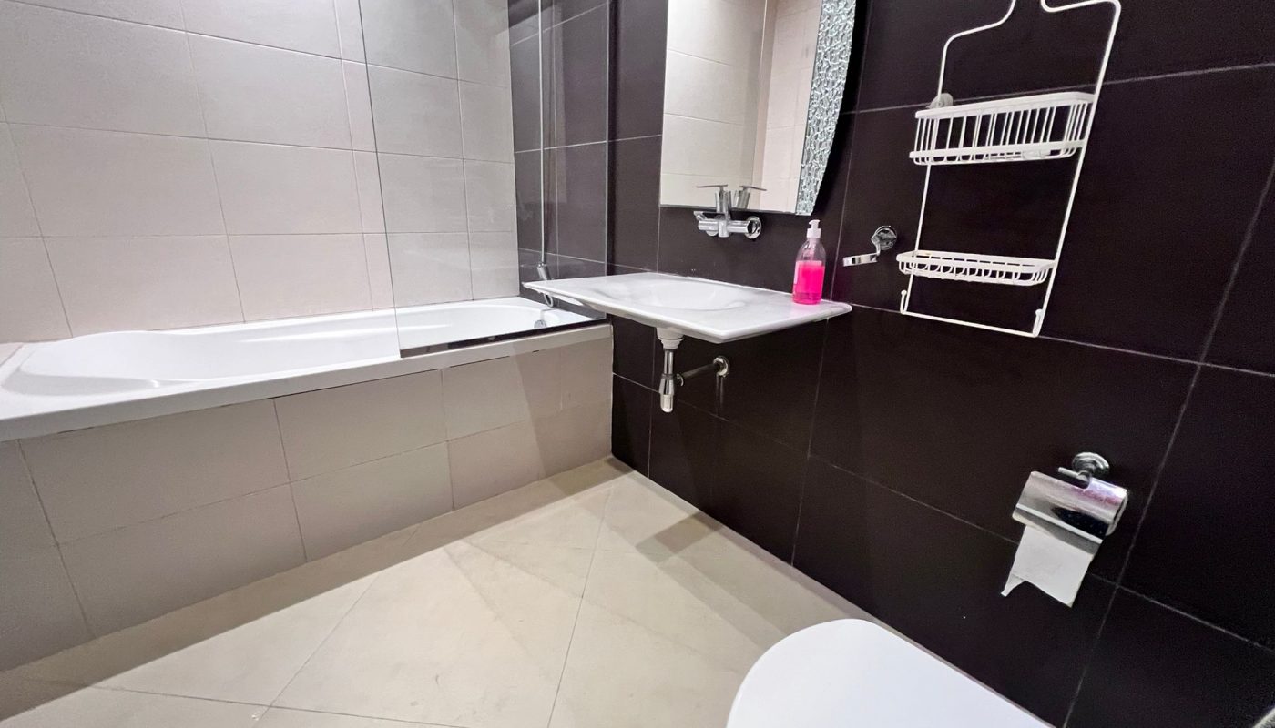 Modern bathroom with a bathtub, wall-mounted sink, mirror, showerhead, wire shelf, toilet, and toilet paper holder; white and dark brown tiles.