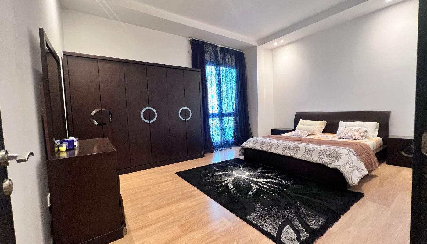 A modern bedroom with a double bed, dark wooden wardrobe, dresser, black patterned rug, and large window with dark curtains.