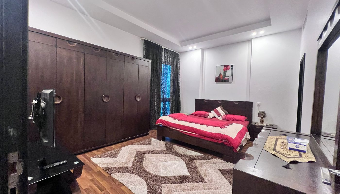 Modern bedroom with a wooden bed, red bedding, large wardrobe, TV, dresser with mirror, patterned rug, and curtains on the window.